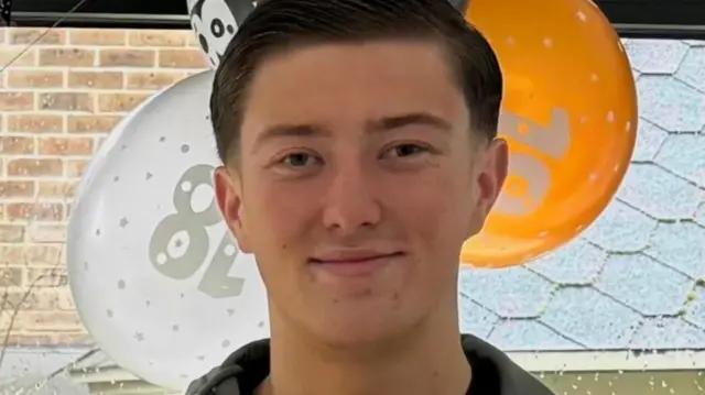 Heny Nowak, 18, standing in front of orange, white and black balloons by a window. He has short dark hair and is smiling and is wearing a blue jacket and a black top underneath.