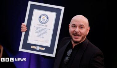 Pitbull wears a dark suit jacket over a dark shirt. He is holding a Guinness World Record award.