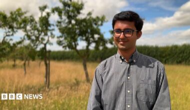 A man wearing glasses and a grey-coloured shirt with open collar. He is standing outdoors in an orchard and is smiling for the camera.