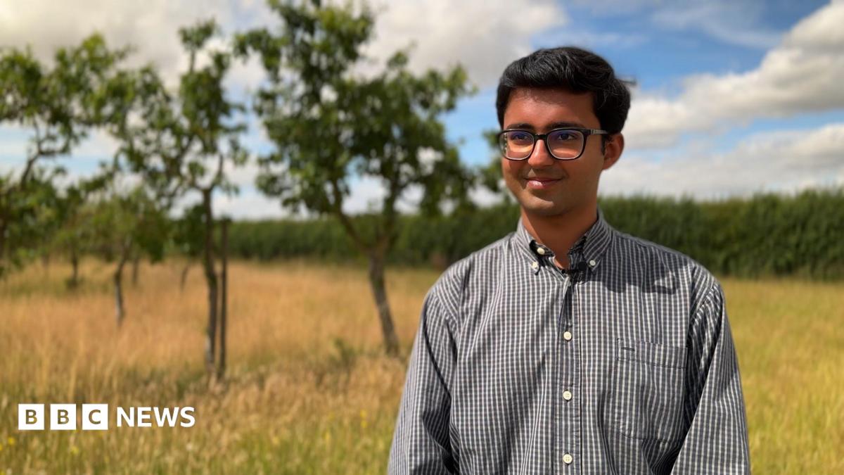 A man wearing glasses and a grey-coloured shirt with open collar. He is standing outdoors in an orchard and is smiling for the camera.