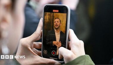 Two hands holding a phone with Zack Polanski on the screen