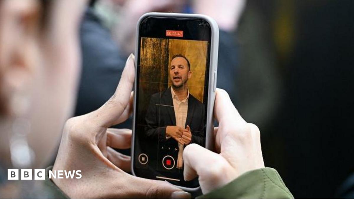 Two hands holding a phone with Zack Polanski on the screen