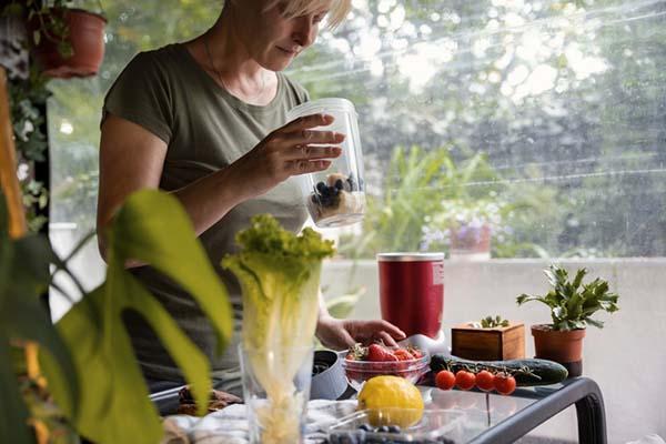 A woman holds a containter with berries and banana slices; there are other fruits and vegetables on a table in front of her.