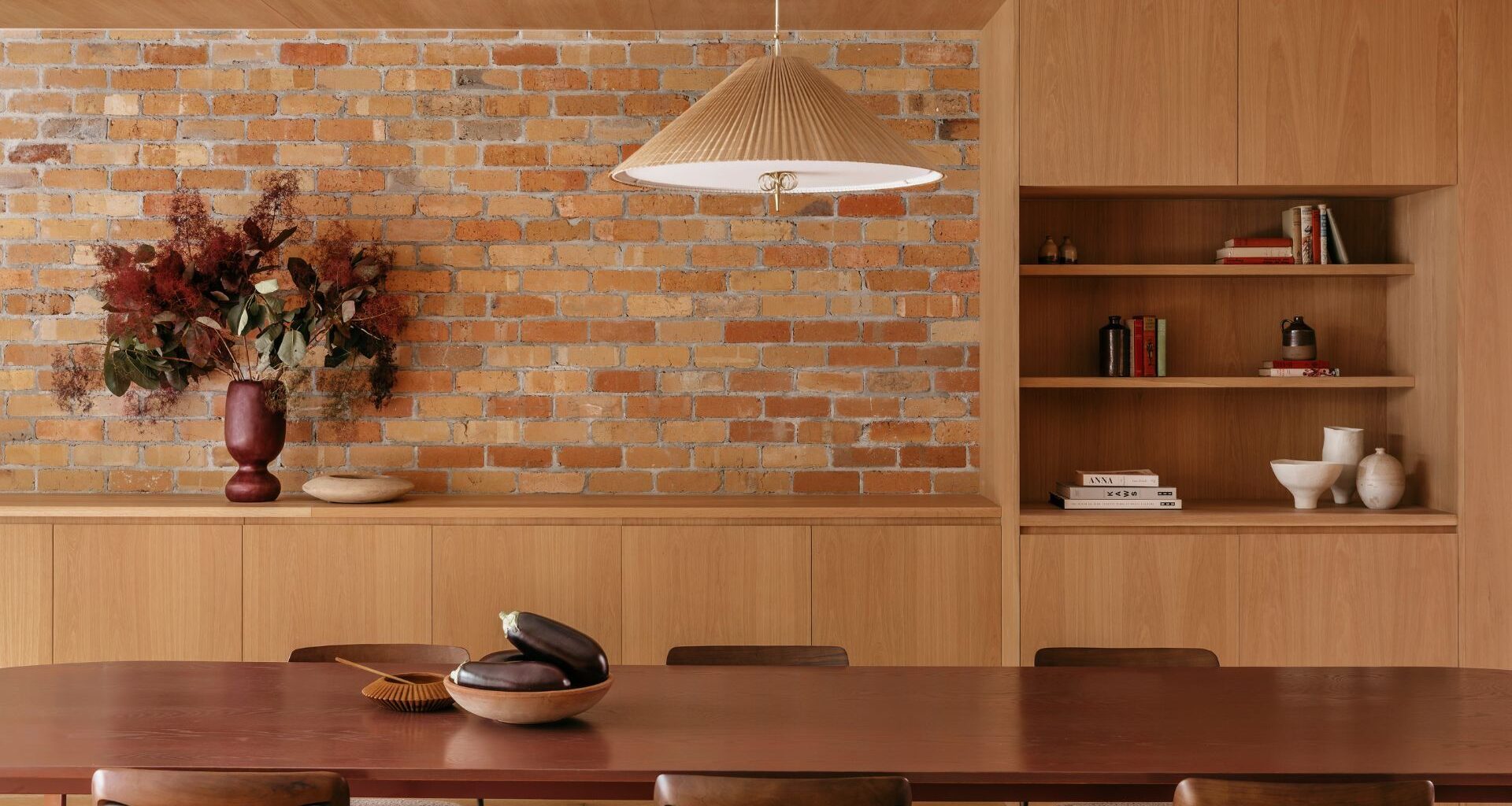 A bright open dining room with a brick wall, a wood cabinet, a woven pendant, and a wood dining set with a bowl of aubergines and a vase of flowers