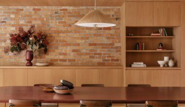 A bright open dining room with a brick wall, a wood cabinet, a woven pendant, and a wood dining set with a bowl of aubergines and a vase of flowers