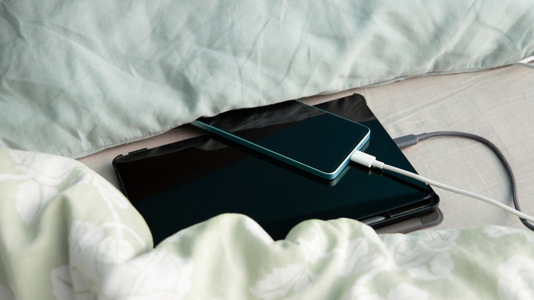 A phone and tablet charging while partly covered by pillows on a bed.