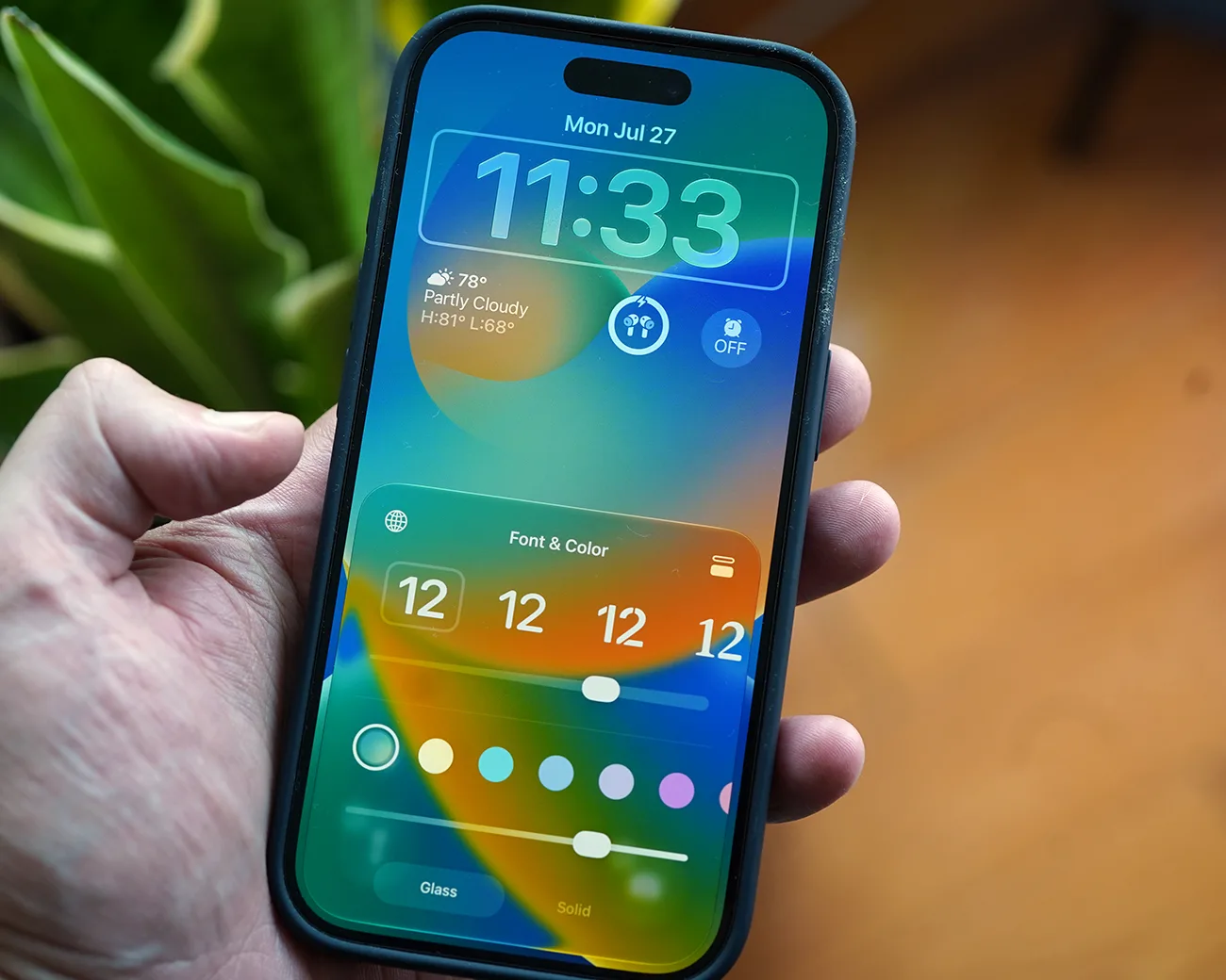 Smartphone with a colorful lock screen showing time, date, weather, and font & color customization options held in a hand.
