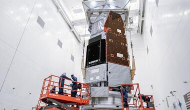 A large silver and gray space telescope sits in a brightly lit, white clean room. Team members in blue suits work on the telescope from orange mechanical lifts.