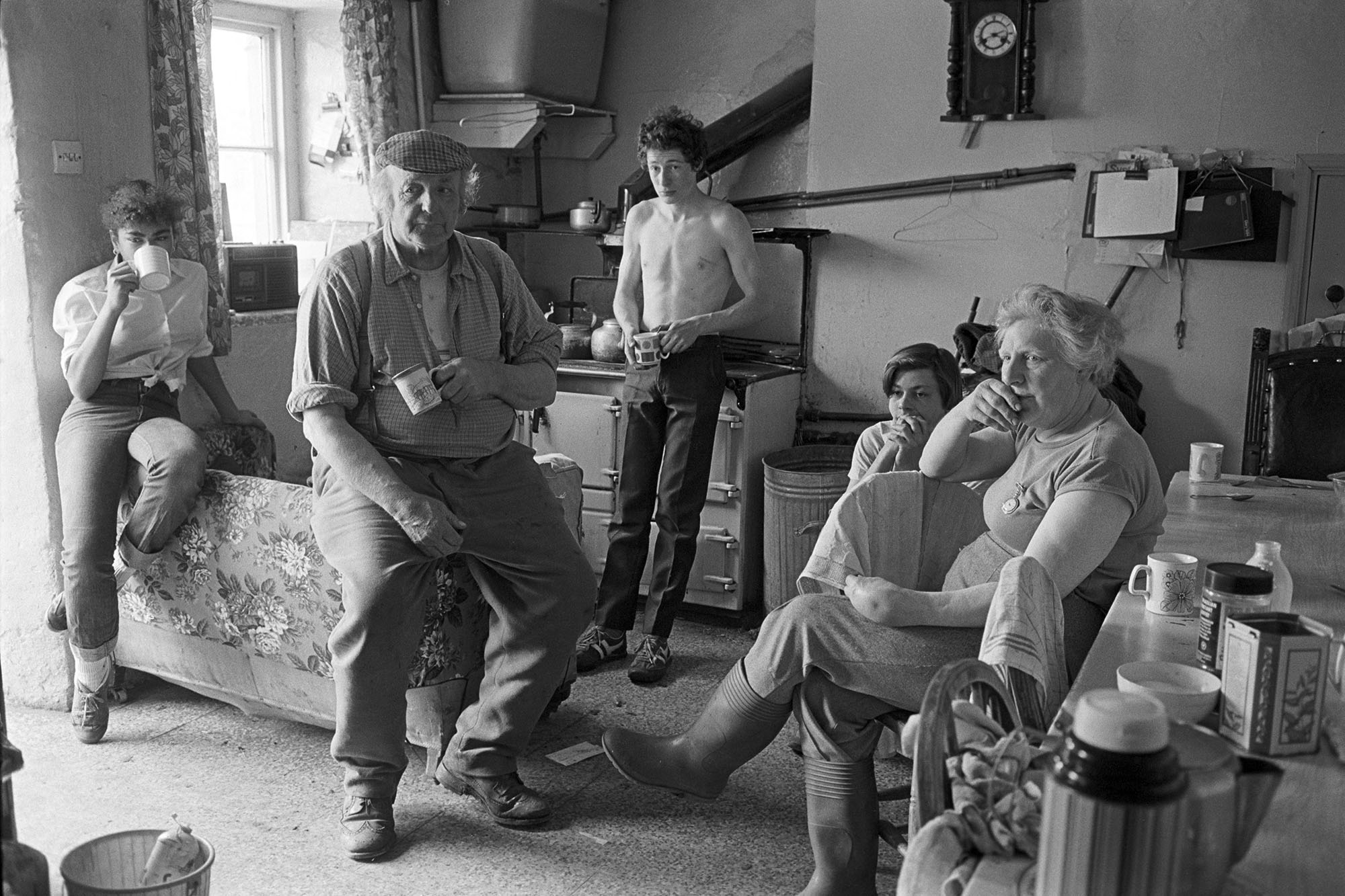 A family gathers in a rustic kitchen holding mugs and looking toward the side as they watch a television broadcast together.