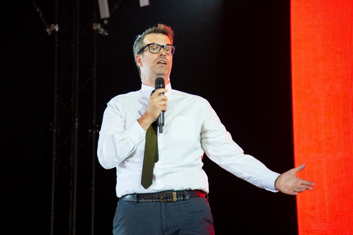 Reading Festival 2022 - Day 2 Marcus Brigstocke speaks into a handheld microphone on stage, wearing glasses, a white shirt and black tie beside a bright red screen.