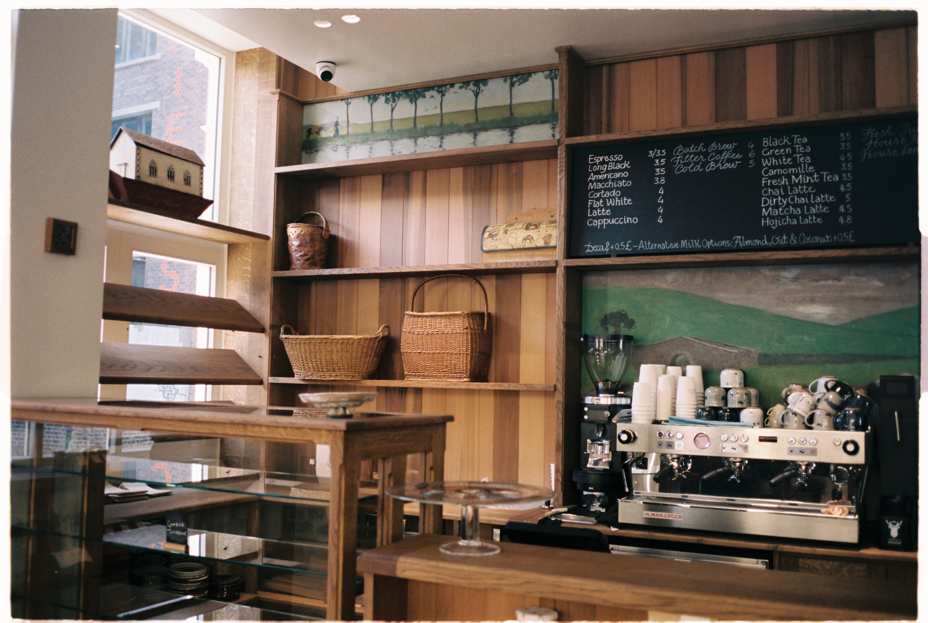 A cafe with modern heritage interiors boasts wooden details and furnishings throughout, including minimal shelving filled with books and collectibles, pastel textiles, plants, and objects throughout.