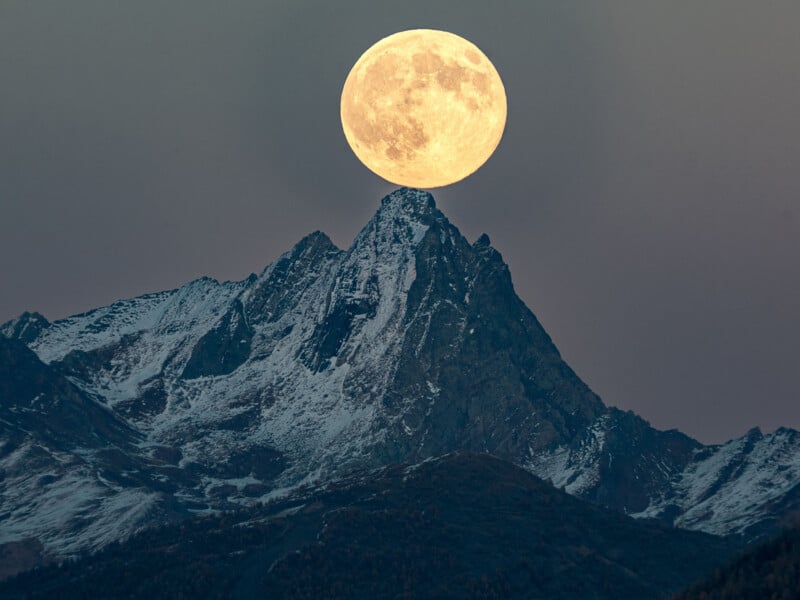 A full moon appears to rest perfectly atop the pointed peak of a rugged, snow-dusted mountain at dusk, creating a dramatic and surreal landscape scene.
