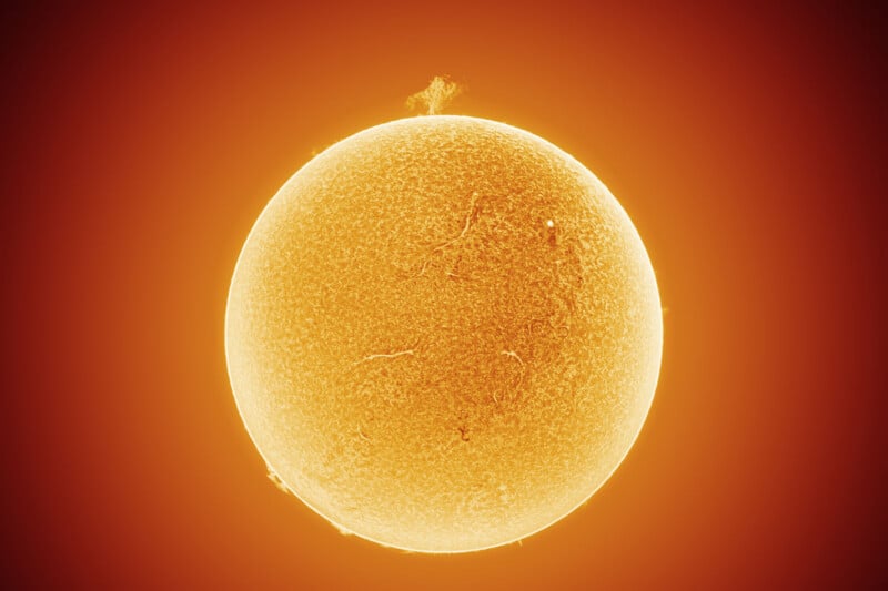 A detailed, close-up image of the Sun shows a glowing orange sphere with textured surface patterns and bright prominences extending from its edges against a dark background.
