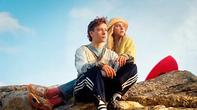 A scene from Silent Roar. Two people sit on a large rock in the sunshine. A blue sky fills the background.