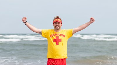 Iain Sterling stood on a beach with his arms out. He is wearing a yellow and red life guard uniform