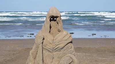 A sand sculpture of a monster with glowing white eyes on a beach with the sea in the background