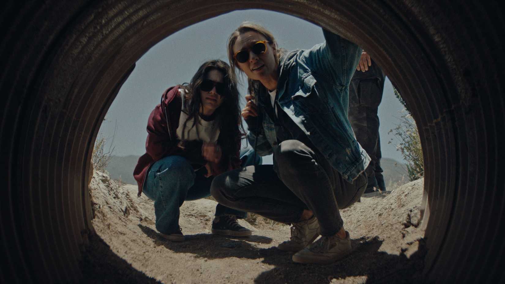 Mia (Stella Marcus) a teenage girl with dark hair wearing sunglasses, and Irene (Michaela McManus), a middle-aged woman also wearing sunglasses, crouch in front of a sketchy-looking tunnel in Redux Redux.