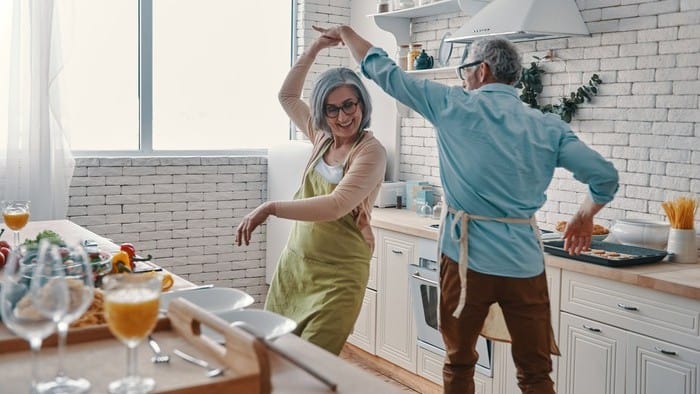 A mature aged couple dance together in their kitchen while they are preparing food in a joyful scene.