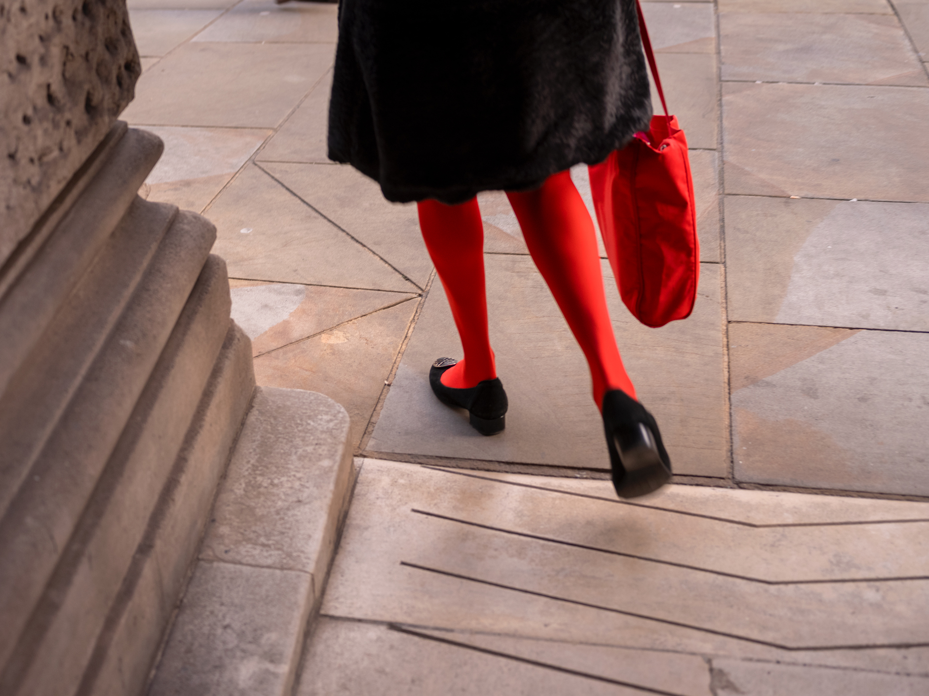 Seen from behind, a pair of bright red tights and black shoes descend a stone step beside an ornate stone column.