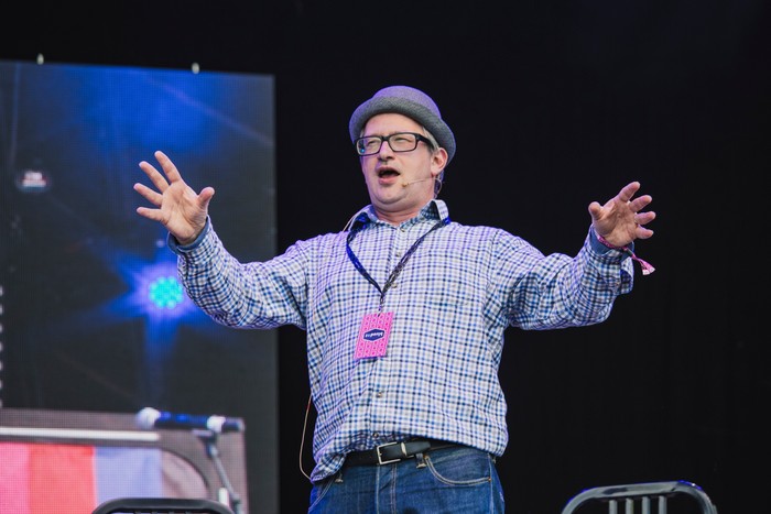 Bluedot Festival 2016 - Day 1 Robin Ince performs on stage in a grey hat, glasses and blue checked shirt, stretching both arms out beneath dark festival lighting.