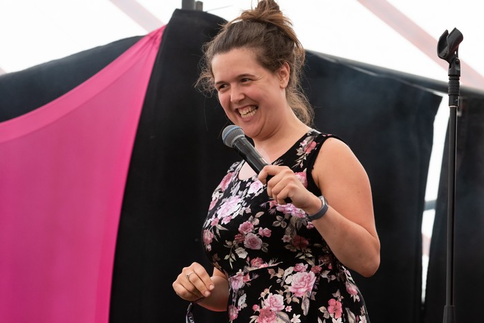 Latitude Festival 2019 Rosie Jones smiles while holding a microphone on stage, wearing a black floral dress against pink-and-black festival draping.