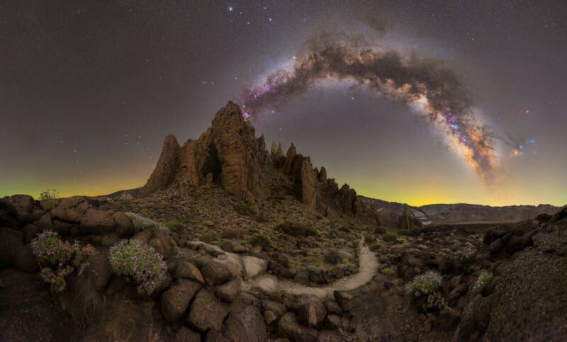 A rocky desert landscape with jagged peaks under a clear night sky, featuring the Milky Way arching brightly overhead. Sparse vegetation and a winding path are visible among the rocks in the foreground.