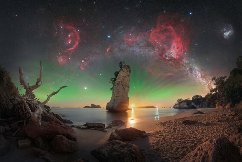 A stunning beach scene at sunset with a tall rock formation in the water, dramatic driftwood on the sand, and a vibrant, star-filled night sky with colorful nebulae and the Milky Way arching overhead.