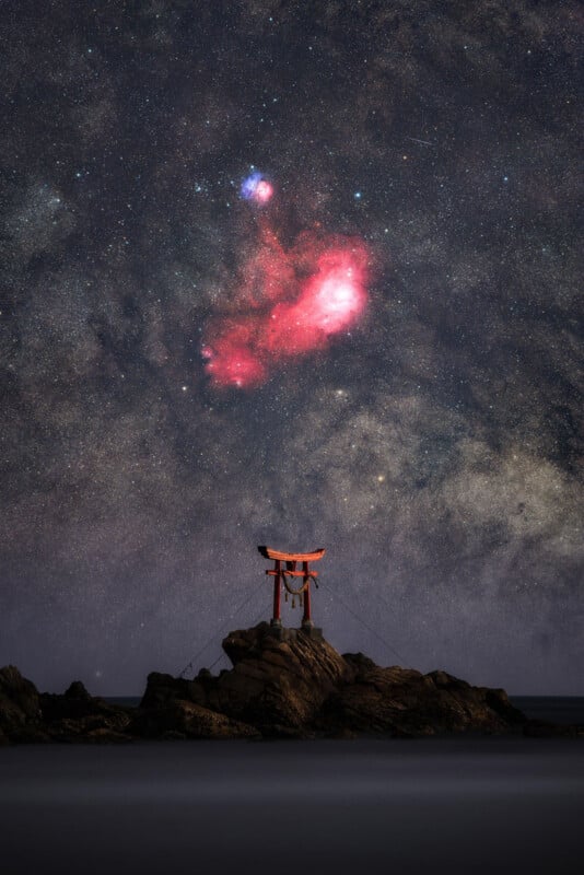 A red torii gate stands on rocky terrain by the sea under a starry night sky, with a vivid red nebula and bright stars visible above.