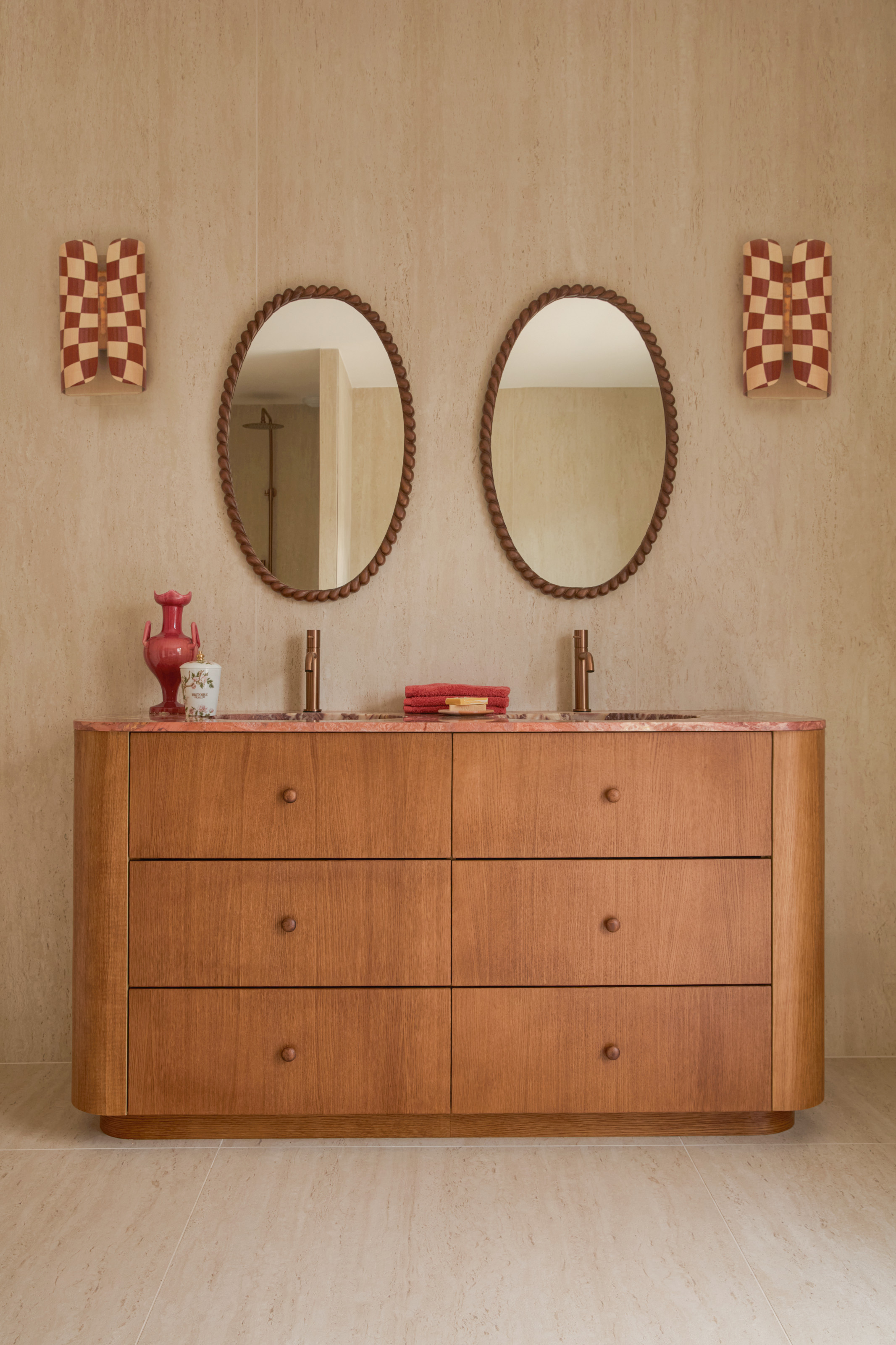 Vanity cabinet in a bathroom with wood finish, curved edges, pink stone countertop, brass taps above twin sinks, twin braided oval mirrors and red and white checkerboard wall lights