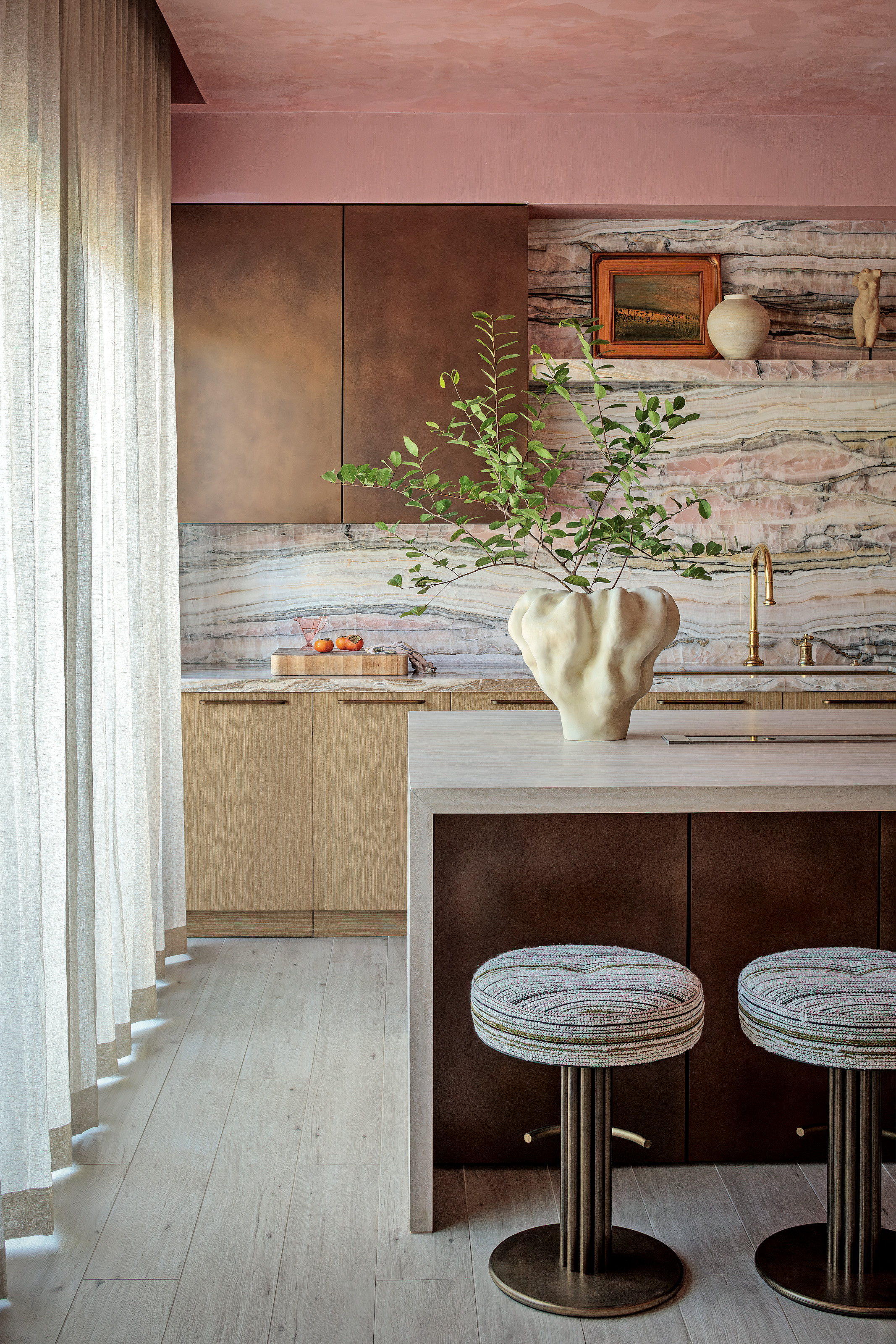 Kitchen with wood and antique brass cabinets, white stone worktop, striped barstools, pink and grey splashback and pink textured ceiling