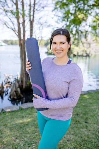woman poses outside with rolled up yoga mat