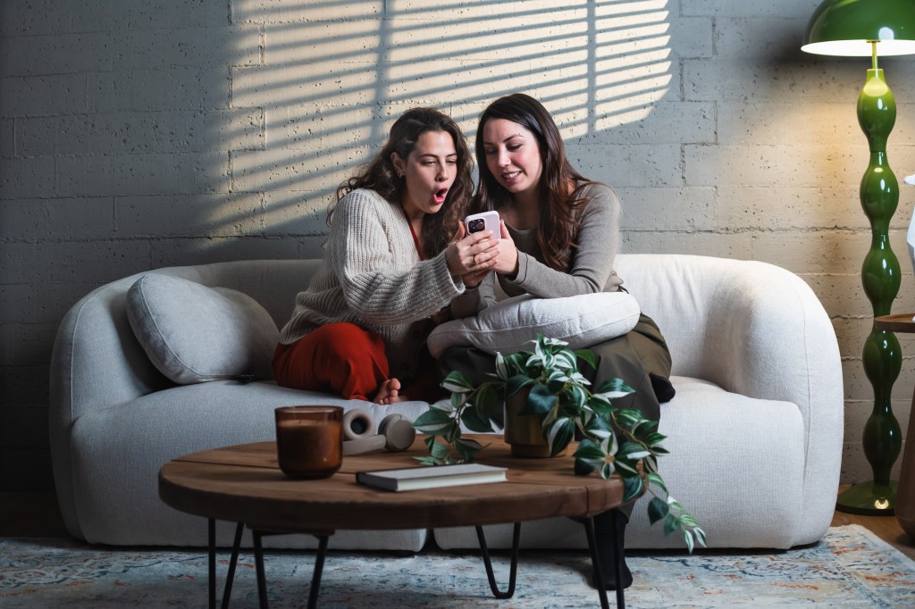 Two women sharing surprising news on a smartphone while sitting on a sofa.