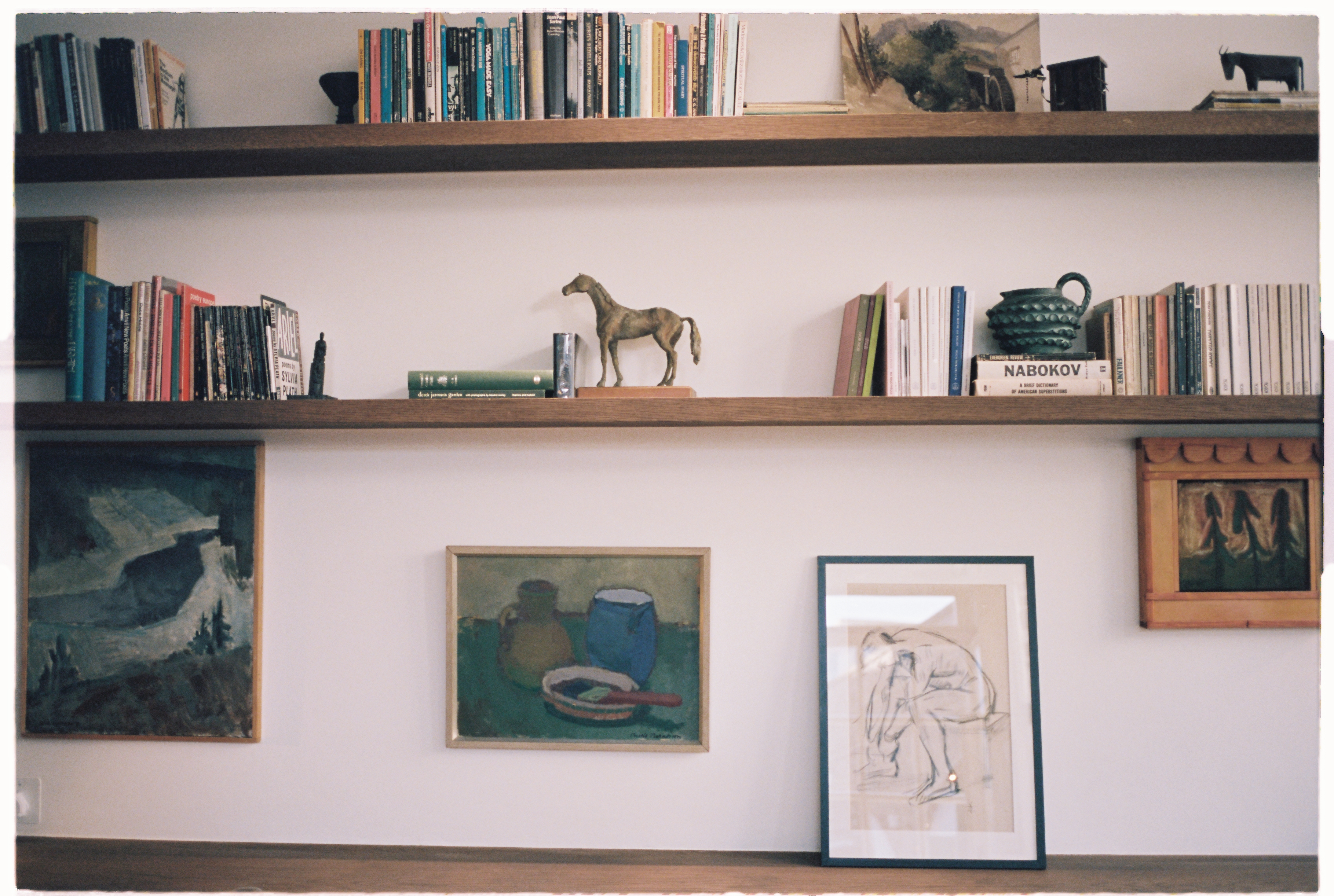 A cafe with modern heritage interiors boasts wooden details and furnishings throughout, including minimal shelving filled with books and collectibles, pastel textiles, plants, and objects throughout.
