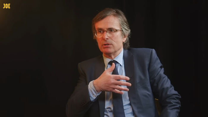 Expert in a suit and glasses discusses an AI warning, gesturing with his hand against a dark background.