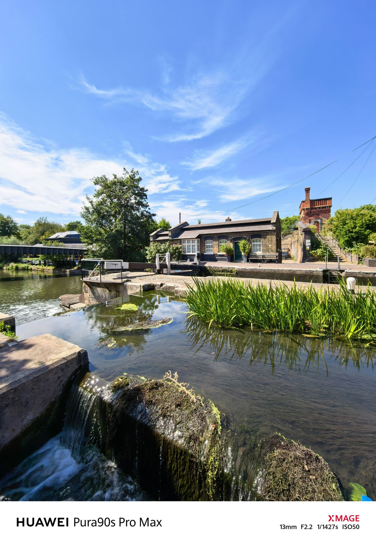 Wide view of a canal lock with flowing water, a lock keeper’s cottage and a red-brick building beyond.