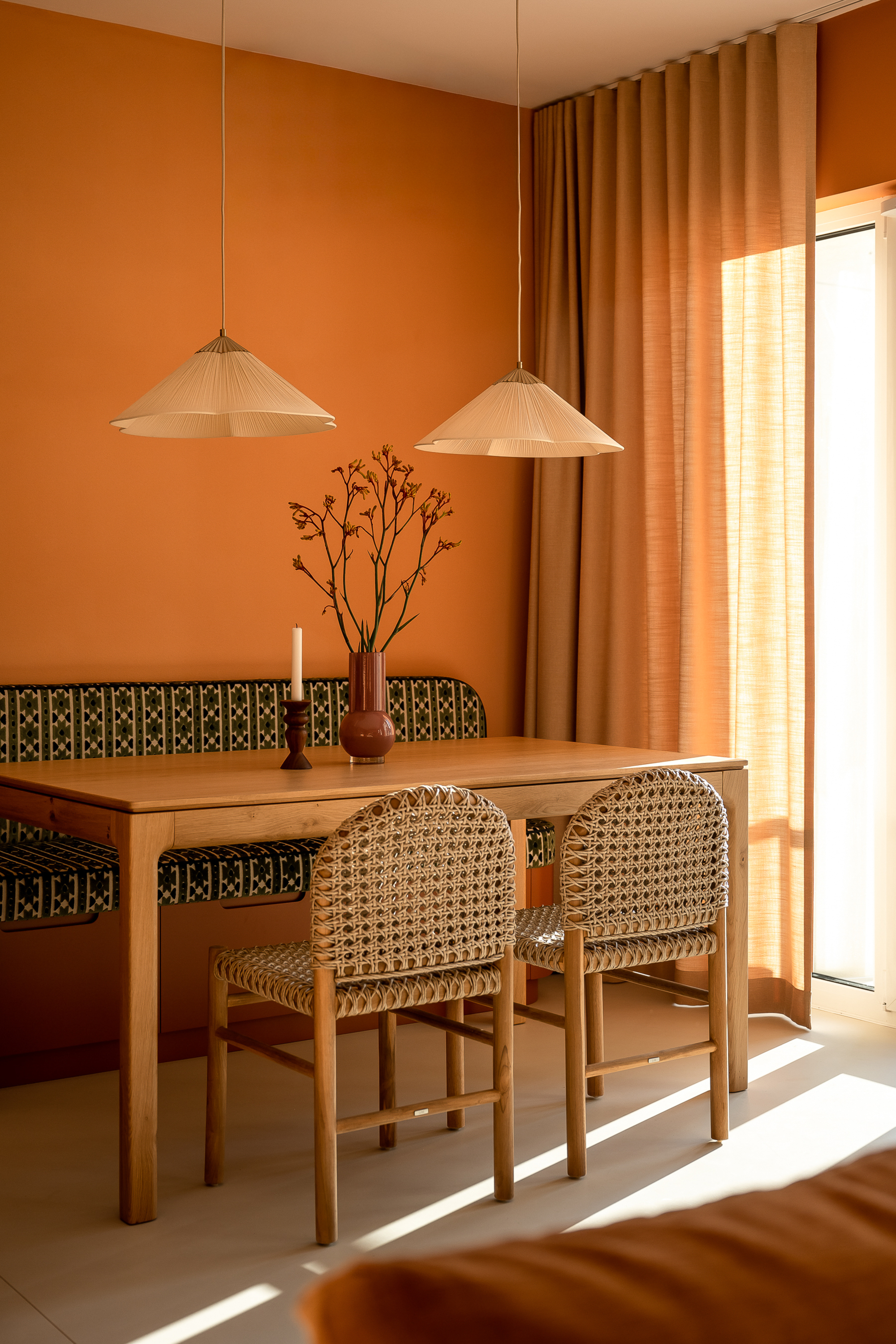 Image of a dining nook painted in a bright orange color. There is a window with tan curtains, a green patterned bench seat, a wooden dining table, and two rattan dining chairs.