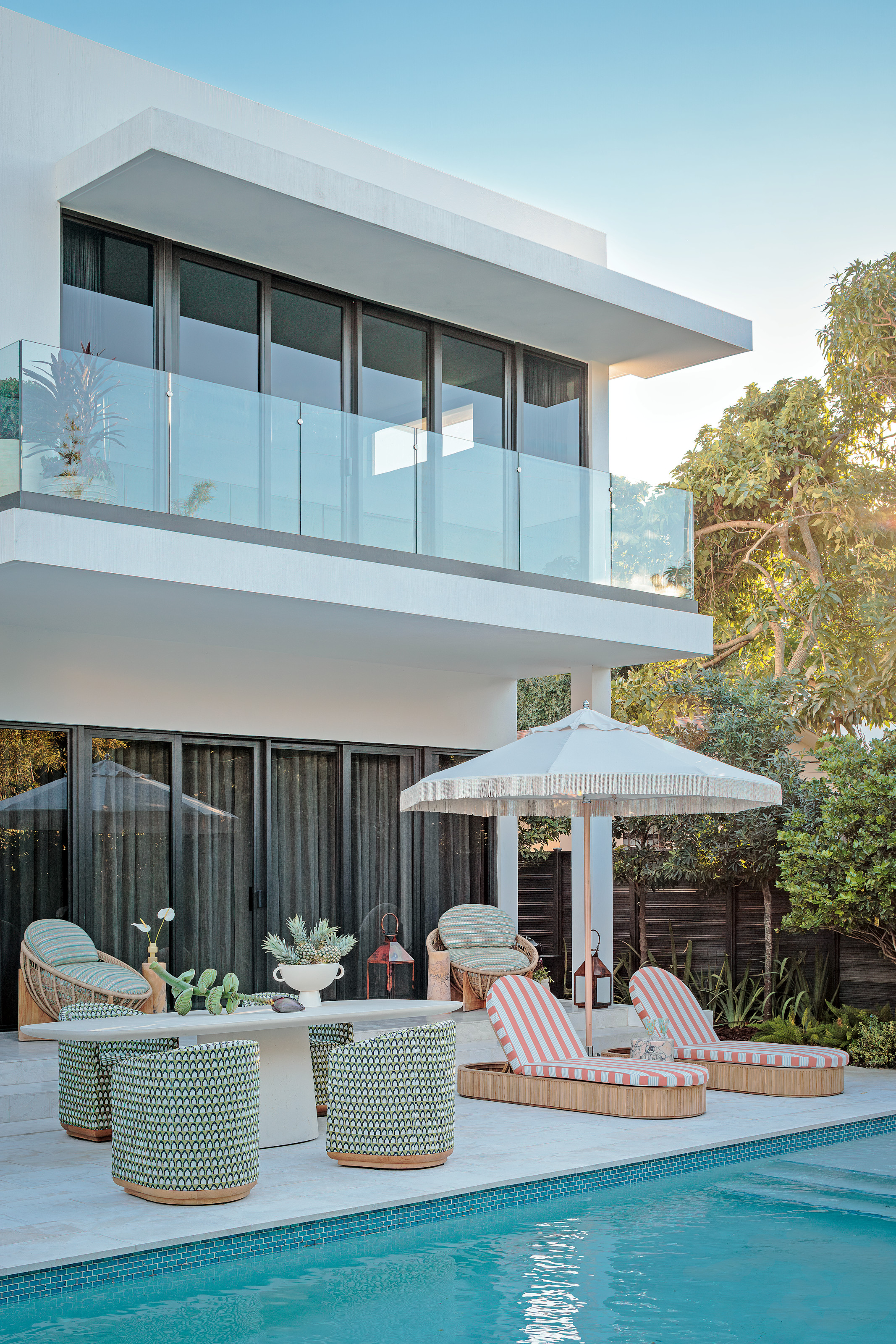 Poolside area with pink and white striped sun loungers, white fringed umbrella, green and white tub armchairs and white oval dining table