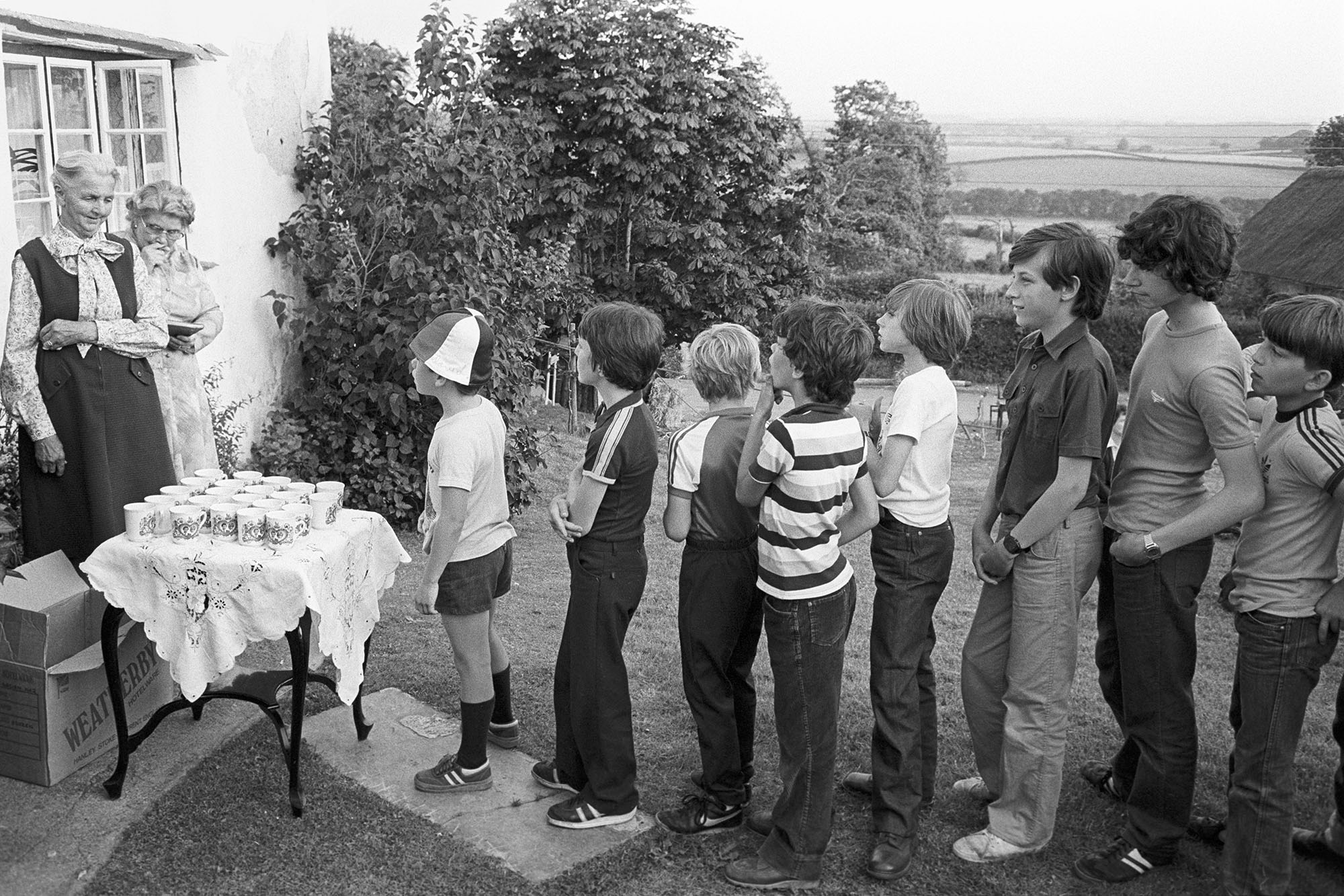 A group of children line up outdoors in a grassy yard to receive commemorative mugs from two elderly women standing next to a small, lace-covered table.