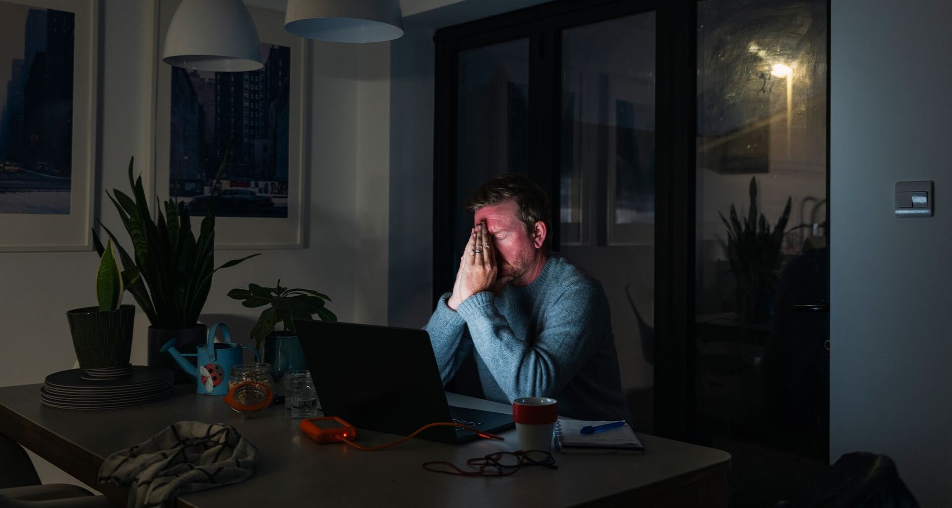 Male office worker sitting at a desk in a dimly lit room with hands held to his face in frustration with light from screen shining on face.