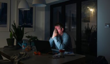 Male office worker sitting at a desk in a dimly lit room with hands held to his face in frustration with light from screen shining on face.