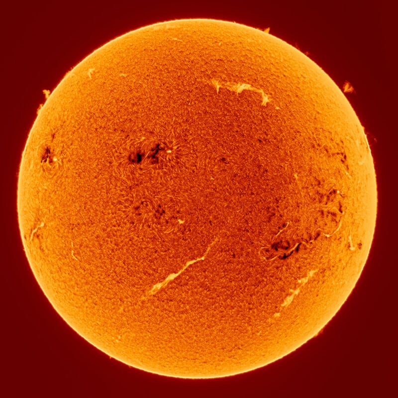 A detailed, close-up image of the Sun showing its fiery orange surface, with visible dark sunspots, swirling textures, and solar prominences along the edges against a deep red background.