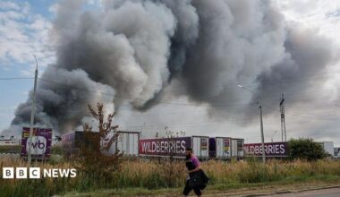 Wildberries employee walks past plumes of smoke rising above wildberries logistics  complex. There are seven company lorries in front of the smoke as well as roadside shrubs