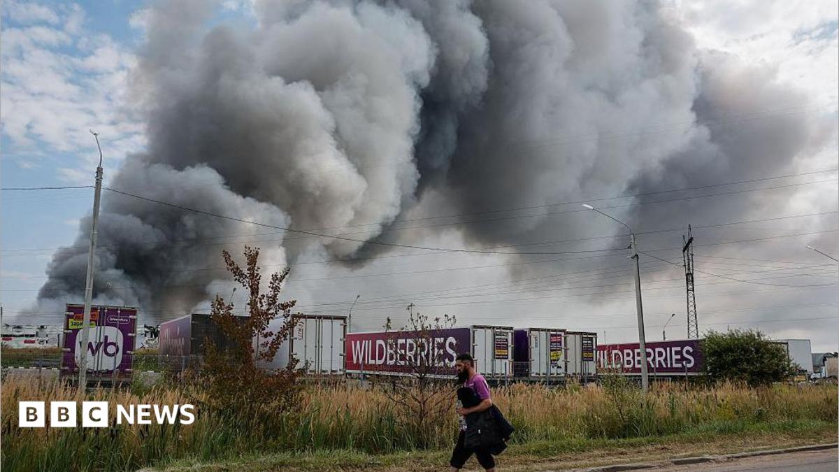 Wildberries employee walks past plumes of smoke rising above wildberries logistics  complex. There are seven company lorries in front of the smoke as well as roadside shrubs