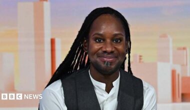 Head shot showing Professor Jason Arday sitting in the Sunday with Laura Kuennsberg studio. He is smiling at the camera with long black locs, wearing a white shirt with a grey waist coat.