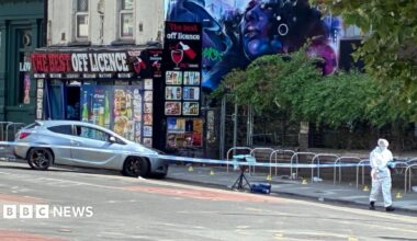 Silver car pushed nose-first against the curb on a road. It is behind a blue and white police tape cordon. Someone in white forensic clothing can be seen in the foreground. An off-licence can be seen in a background, with a dystopian blue-hued mural of a woman on the side.