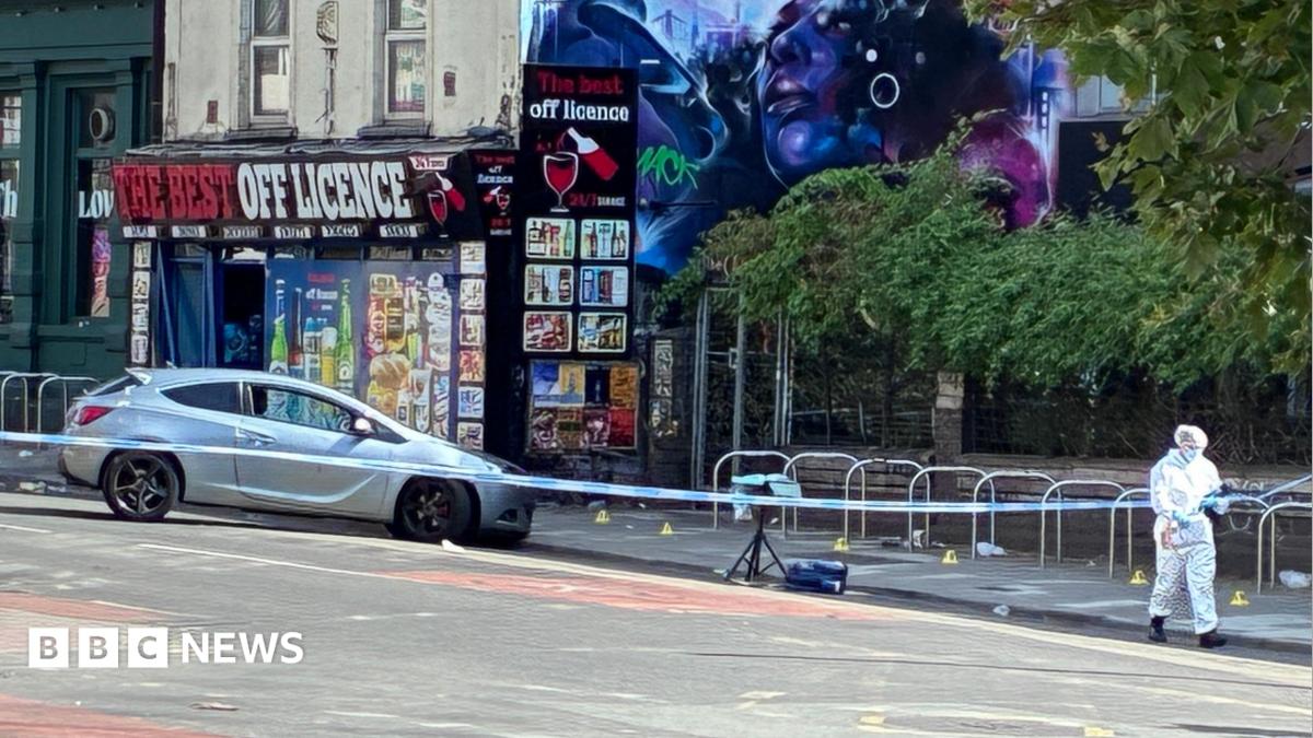 Silver car pushed nose-first against the curb on a road. It is behind a blue and white police tape cordon. Someone in white forensic clothing can be seen in the foreground. An off-licence can be seen in a background, with a dystopian blue-hued mural of a woman on the side.