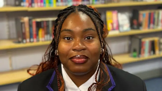 Gift has long brown dreadlocks and smiles at the camera. She wears a black school blazer and white shirt and appears to be in a library
