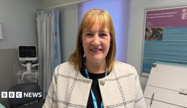 A woman wearing a light-coloured checked jacket and an NHS lanyard, standing in a hospital room. Medical equipment, a curtained area and information boards are in the background.