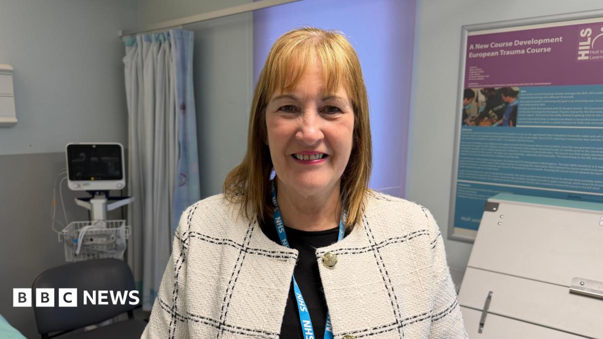 A woman wearing a light-coloured checked jacket and an NHS lanyard, standing in a hospital room. Medical equipment, a curtained area and information boards are in the background.