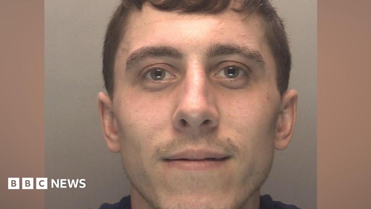 A police mugshot of a man in his early 20s, who has brown hair and light stubble. He is smiling at the camera.